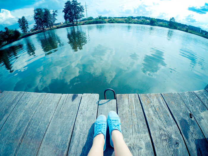 A girl in blue shoes sits on an old shabby wooden pier near the lake river in a summer sunny day