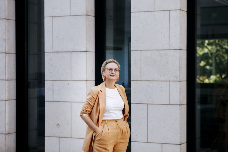 Businesswoman with hands in pockets looking away while standing against wall