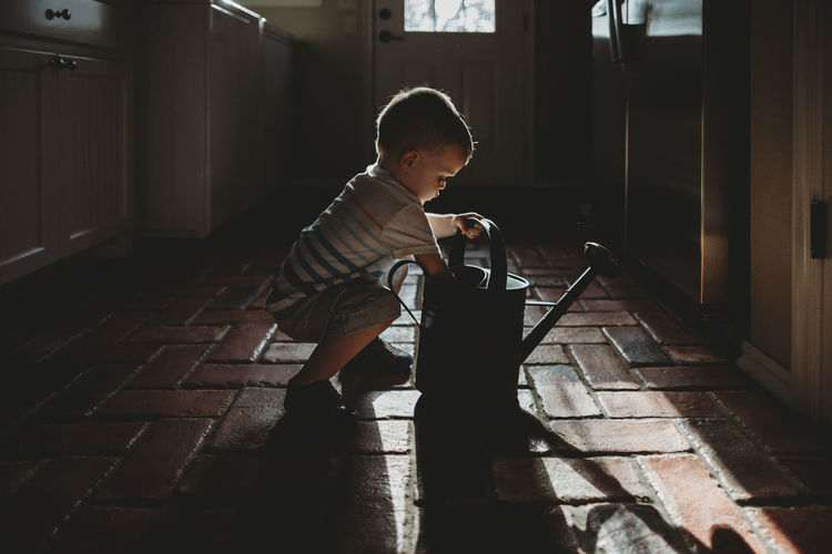 Side view of baby boy holding watering can while crouching on floor at home