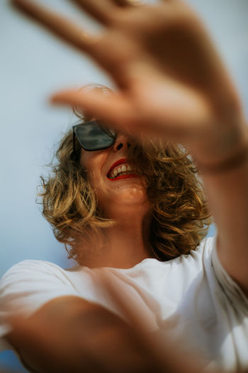 From below stylish brown curly haired woman with red lipstick in trendy sunglasses looking at camera with blue sky on the background