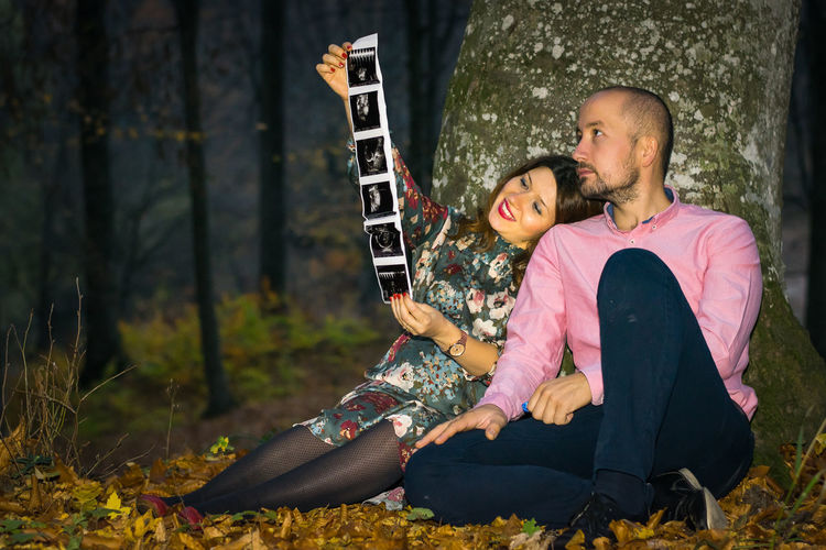Young couple holding ultrasound images while sitting against tree in forest