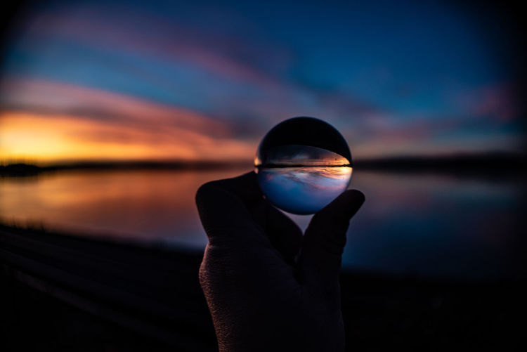 Cropped hand of person holding crystal ball against sky during sunset