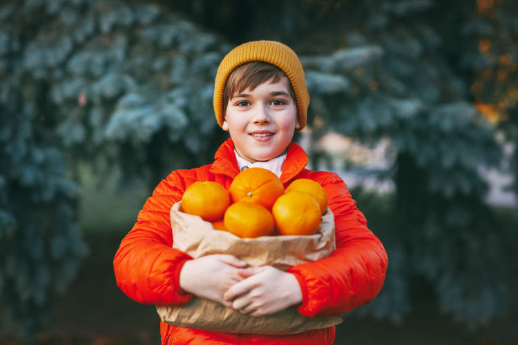 A boy in a bright orange jacket and a yellow hat holds a large package with oranges in his hands 