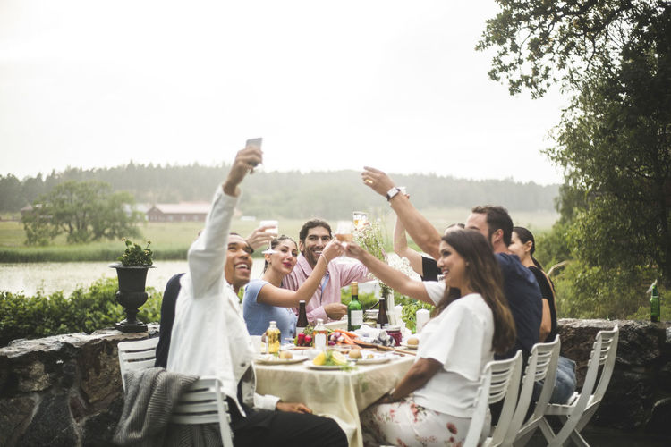Smiling man taking selfie with friends holding drinks during dinner party in backyard