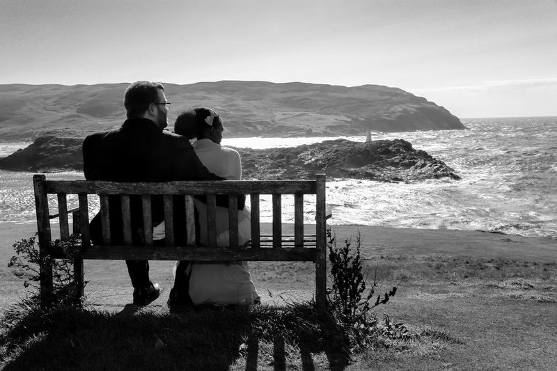 Newlyweds sit on bench overlooking the sea on the isle of man
