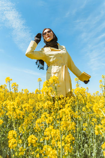 A young stylish woman poses among a blooming yellow field of rapeseed under a bright spring sky