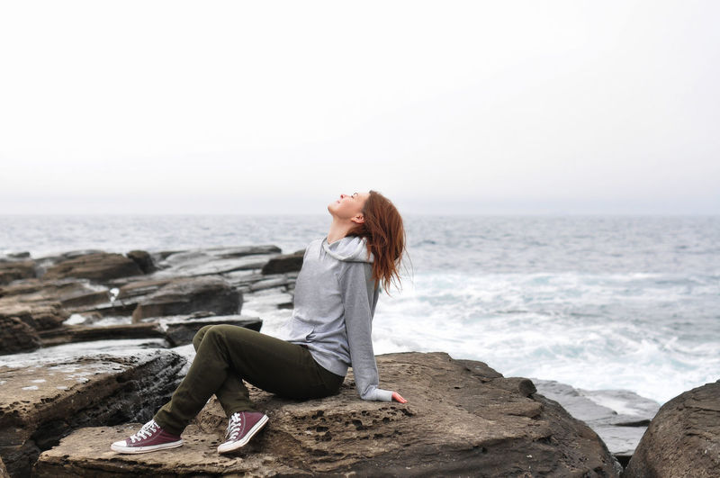 Young woman in hoodie, jeans sits on rocks near the stormy oceans with waves. local tourism