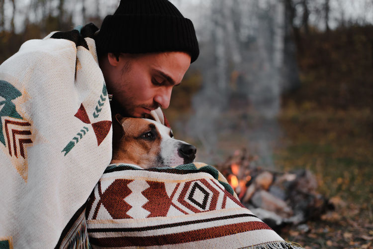 Man in poncho blanket by the camp fire hugs his dog, chilly autu