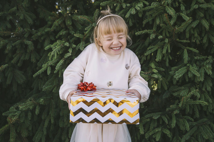 Excited girl holding gift box in front of fir tree