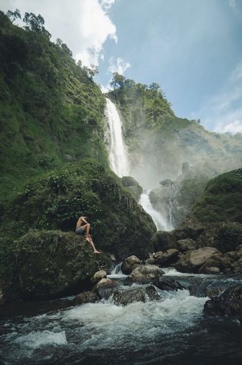 Side view of shirtless man sitting on rock against waterfall