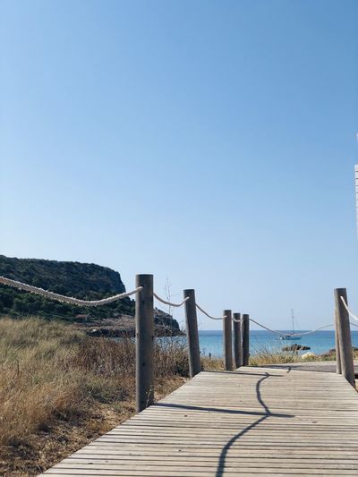 Wooden walkway leading towards sea against clear blue sky