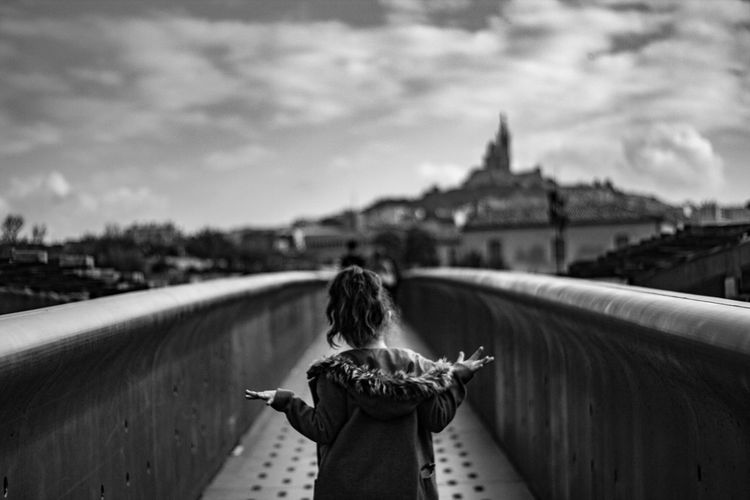 Rear view girl looking at city buildings while standing on footbridge against sky