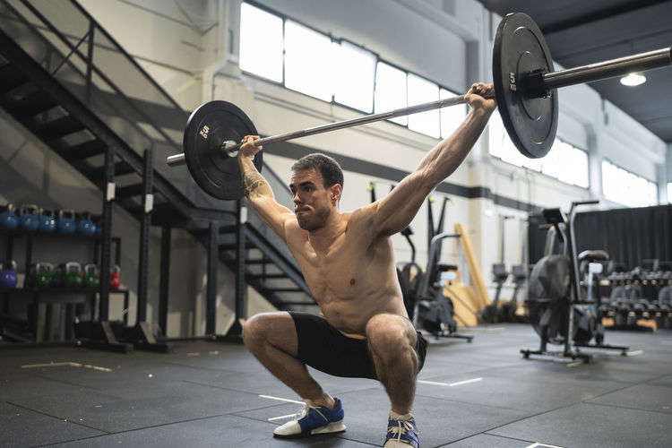 Sportsman crouching while picking up barbell in gym
