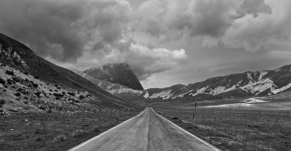 Empty road on landscape by mountains against cloudy sky
