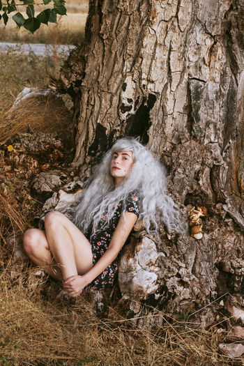 Man in dress and gray wig sitting by tree in forest
