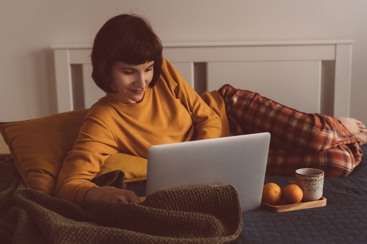 Smiling woman using laptop while sitting on bed at home
