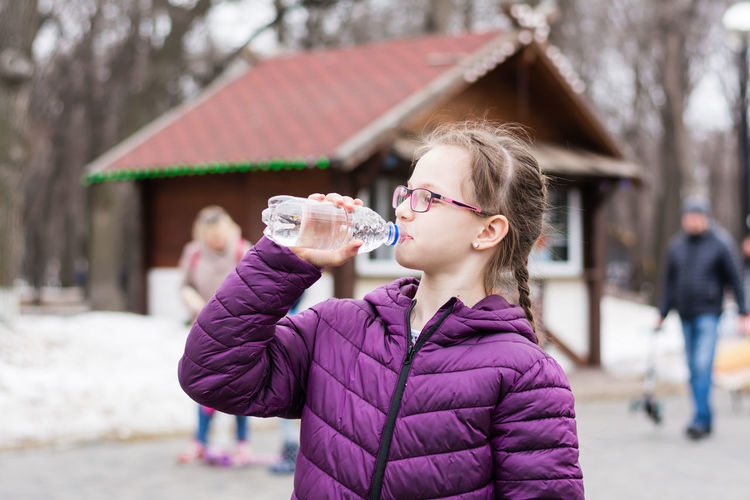 A cute girl in glasses drinks water from a bottle bought in a food truck in a city park
