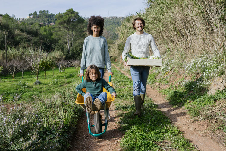 Family walking on a dirt track, pushing wheelbarrow, carrying crate with vegetables