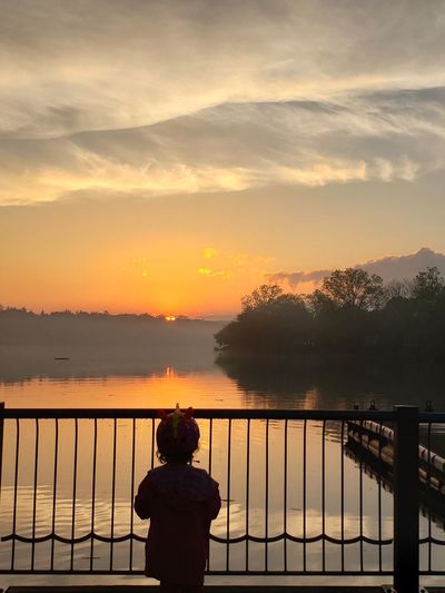 Rear view of girl standing by railing against lake during sunset