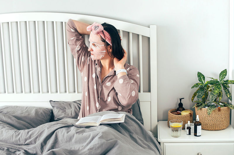 Woman with face mask reading book while while sitting on bed at home