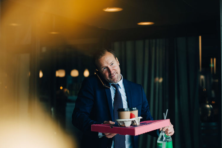 Mature businessman talking on mobile phone while carrying food and drinks in cafe