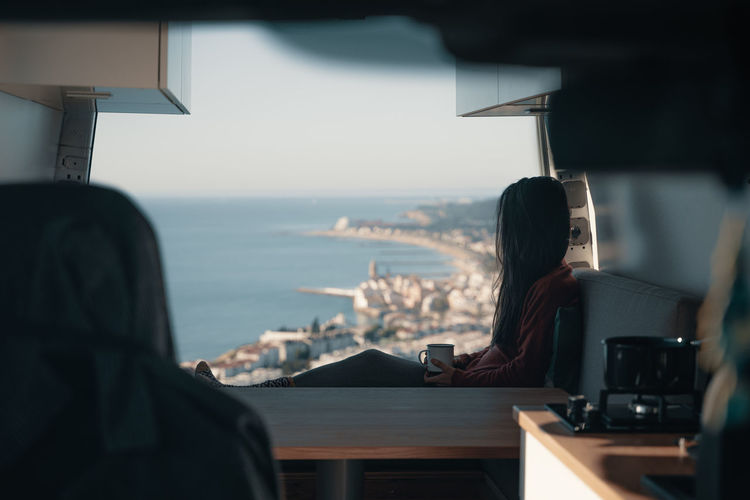 Rear view of woman sitting at table in bus by sea against sky