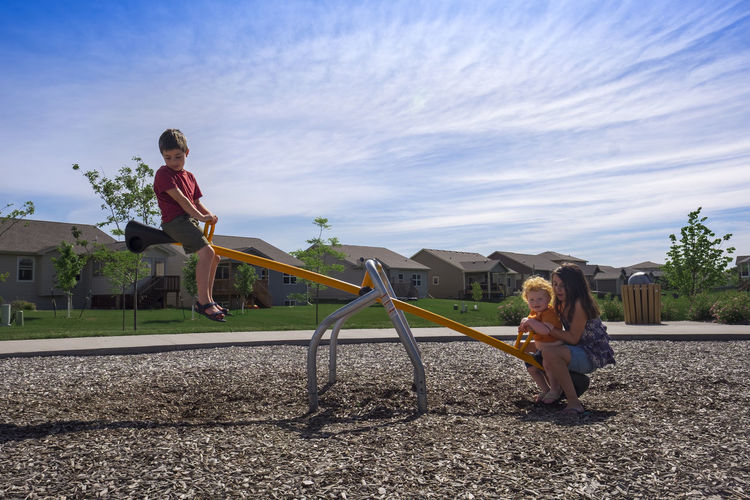 Siblings playing on seesaw against sky at park during sunny day