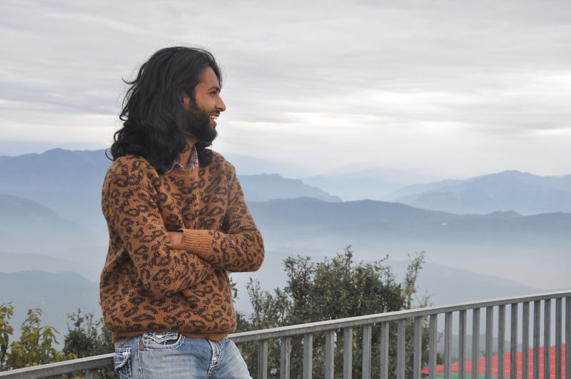 A smiling long haired young man looking sideways with crossed arms while standing in hilly area 