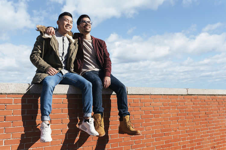 Low angle view of smiling friends sitting on brick wall
