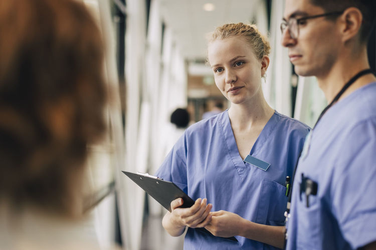 Young female hospital staff holding clipboard while discussing with colleagues