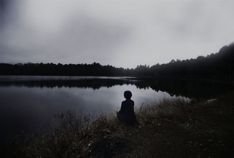 Silhouette man sitting by lake against sky