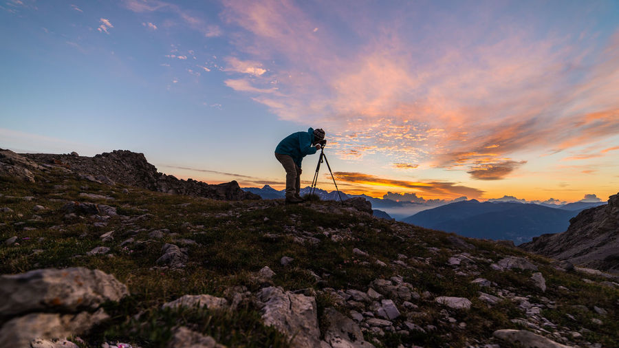 Side view of man photographing on mountain against sky during sunset