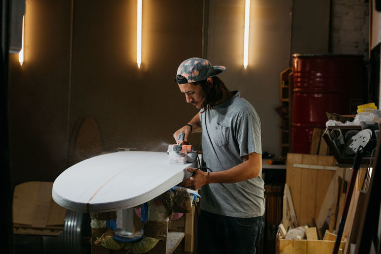 Male shaper using electric planer and polishing surface of surfboard in workshop