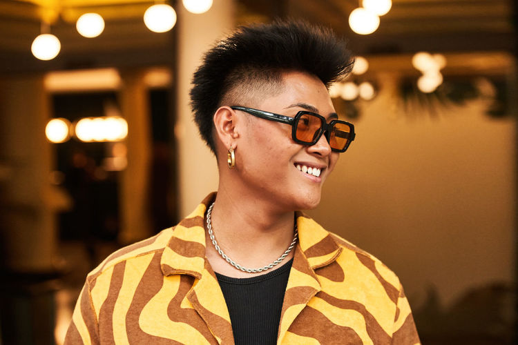 Smiling young non binary male in stylish shirt with sunglasses and accessories with face skin imperfections looking away