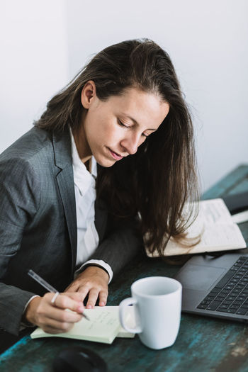 Side view of focused young female entrepreneur in formal wear sitting at table with laptop and cup of coffee and writing information in notebook while planning working day at workplace at home