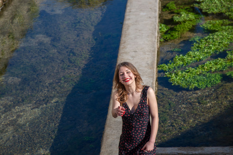 Smiling young woman pointing while standing by canal on retaining wall