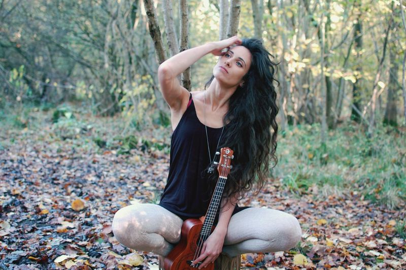 Thoughtful young woman with guitar sitting in forest