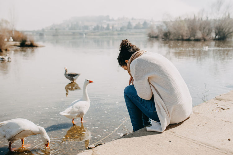 Woman sitting by lake with duck against sky