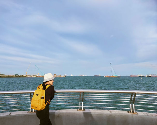 A woman carrying yellow backpack looking at a calm blue ocean on sunny day