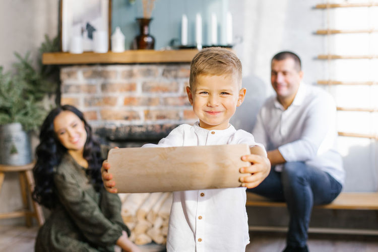 Little boy holds a log in his hands to light a fireplace against the background of happy parents