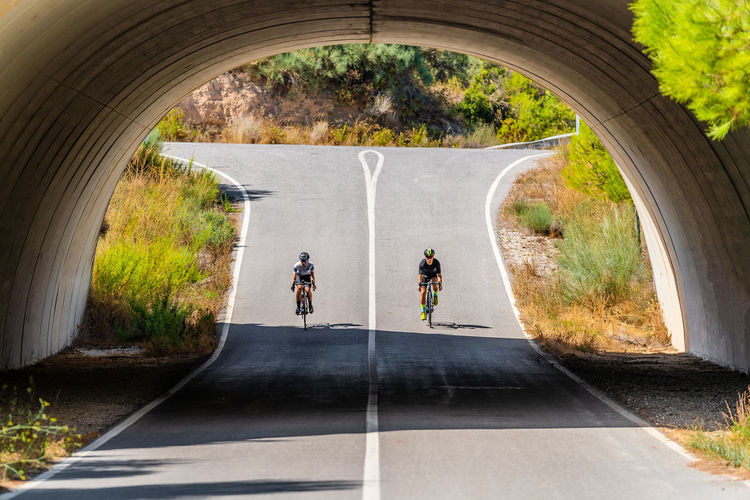 Full length of distant unrecognizable bicyclists in sportswear and helmets cycling together on asphalt roadway under arched bridge in summer countryside
