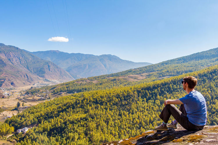 Mid adult man looking at mountains while sitting on cliff against sky