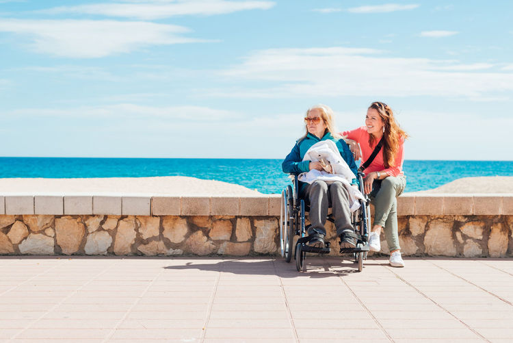 Cheerful adult daughter with elderly mother in wheelchair sitting stone fence along promenade near sea in summer looking away