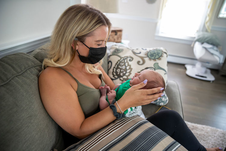 Blonde woman wearing face mask holding sleepy newborn baby.