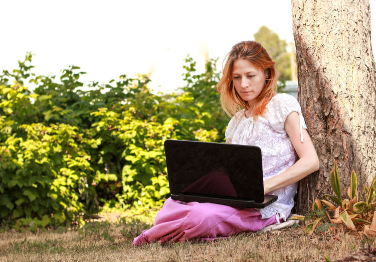 Girl with red hair sits with a laptop in a park near a tree, concept of freelancing,online shopping