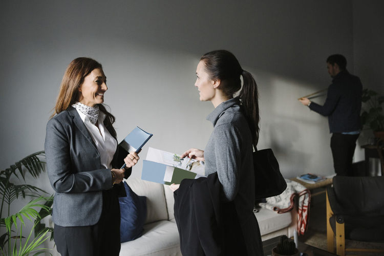 Happy woman and female realtor discussing brochure while man measuring wall in background