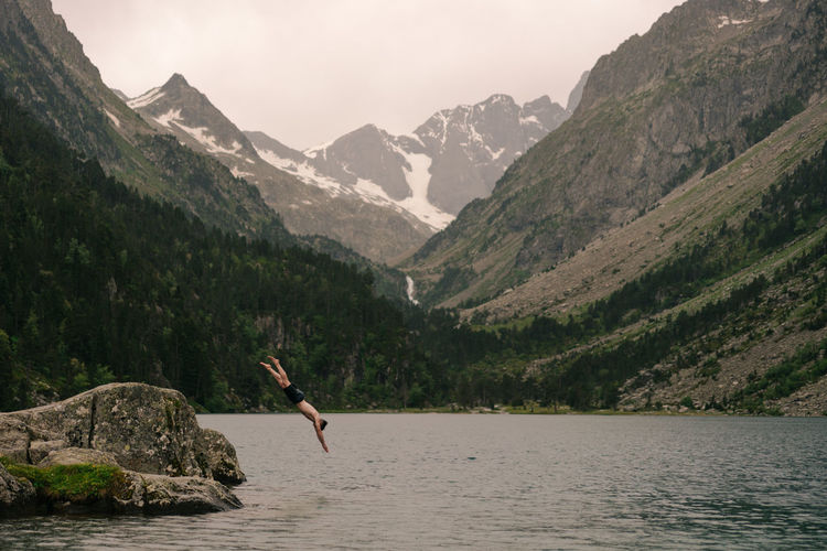 Side view of unrecognizable shirtless male traveler jumping into water of lake surrounded by massive rocky mountains on cloudy day
