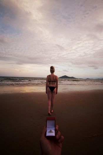 Rear view of woman standing at beach with man holding ring against sky