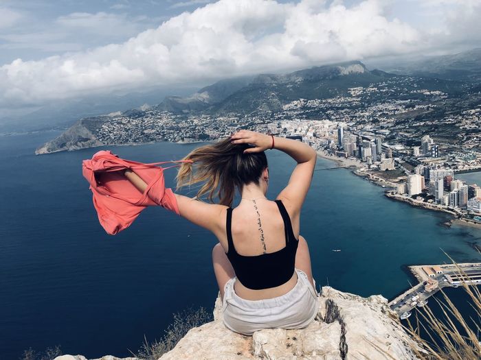 Rear view of woman looking at cityscape sitting at cliff against sky