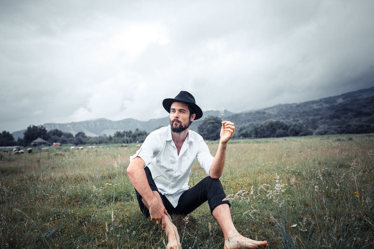 Full length of young man sitting on grassy field against cloudy sky